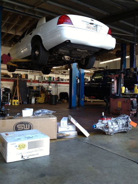 2004 Ford Crown Victoria - A white 2004 Ford Crown Victoria is shown on a lift in a garage, with a new transmission and parts on the floor.