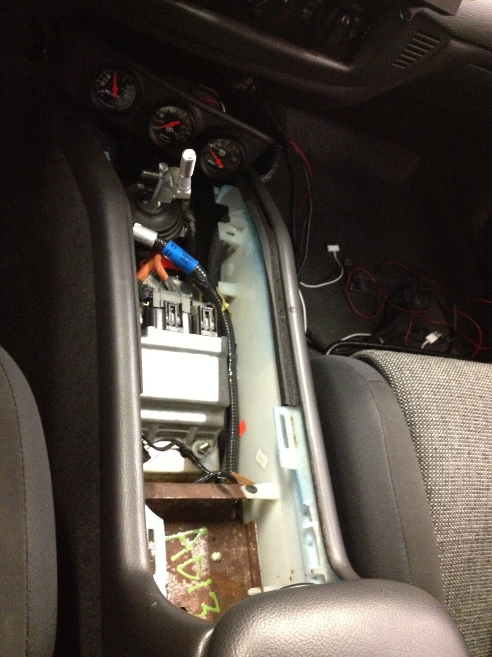 Car interior showing center console removed, exposing wiring, a fuse box, and three gauges on the dashboard