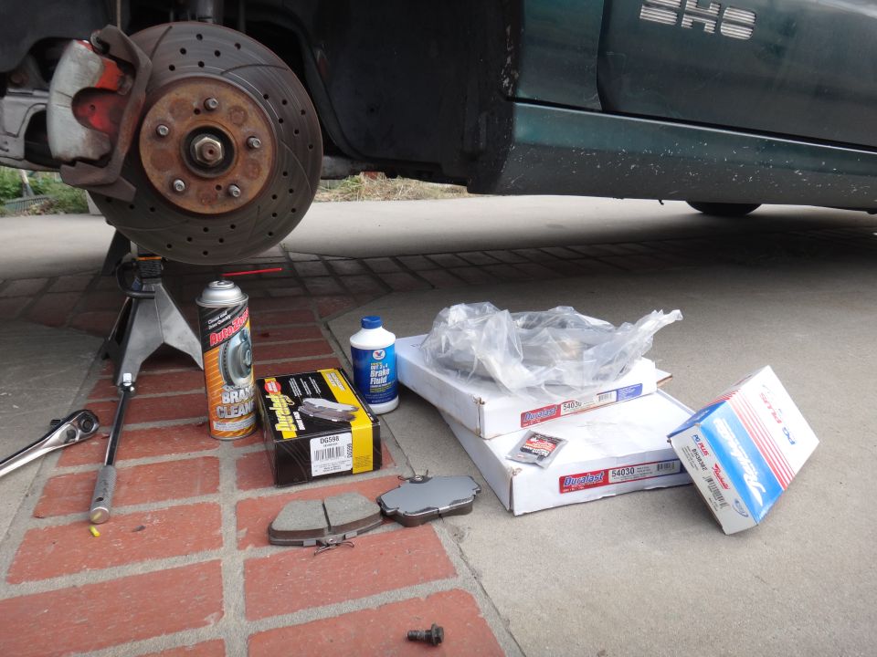 Brake pads, rotors, brake fluid, and brake cleaner spray laid out on the ground next to a car's wheel assembly on a jack stand
