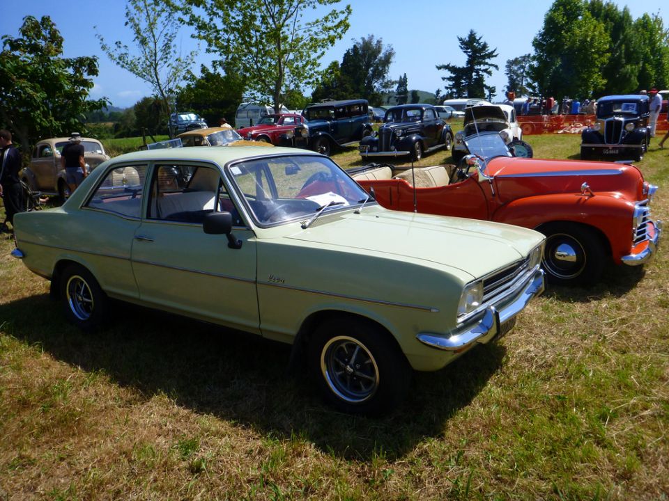 light green sedan parked on grass next to an orange convertible, with other cars and people in background
