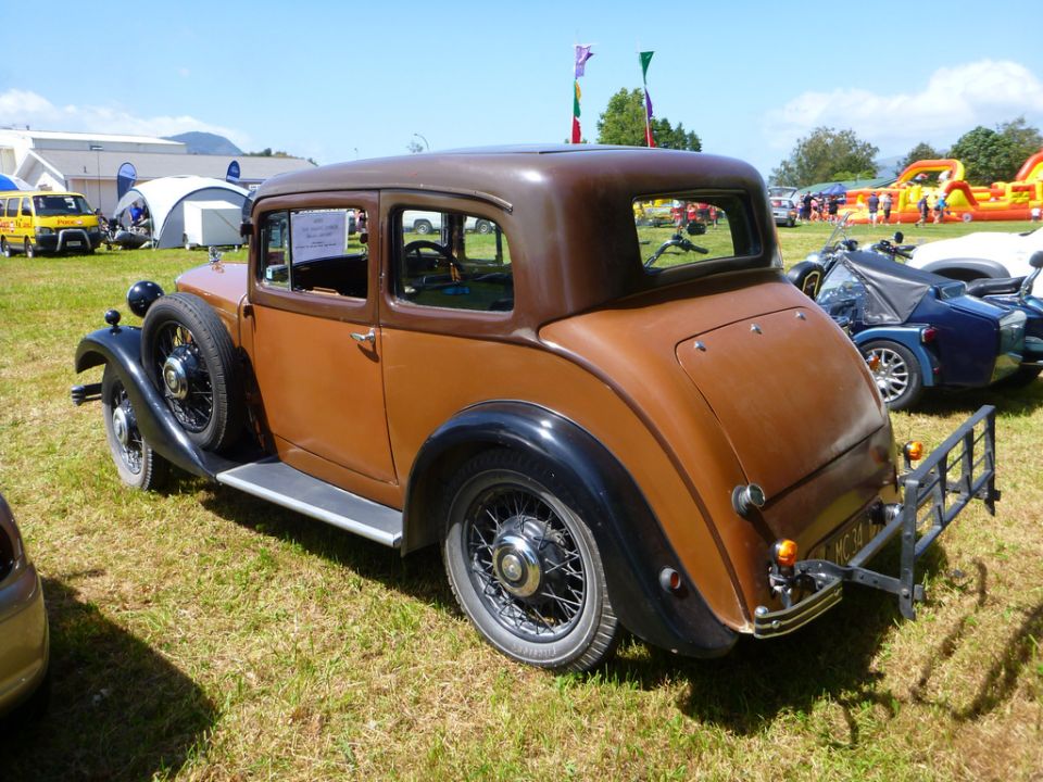 Rear view of a brown vintage sedan with a spare tire mounted on the left front fender. Other vehicles and tents are visible in the background