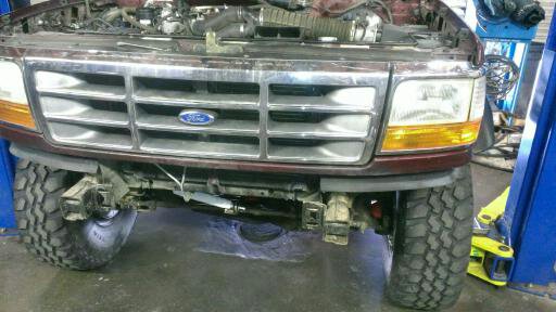 Front of a maroon Ford truck with hood open, showing grille and headlights. Tires and suspension visible