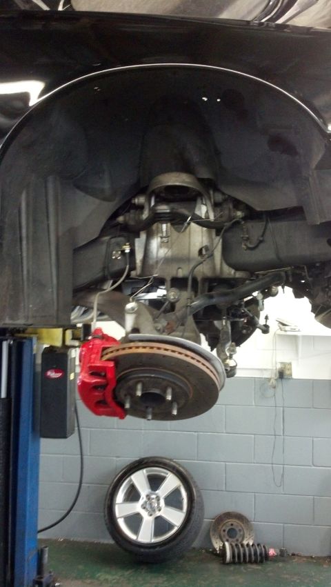 2003 Ford Crown Victoria - vehicle raised on a shop lift showing front suspension, a drilled rotor with red caliper, and a removed alloy wheel below.