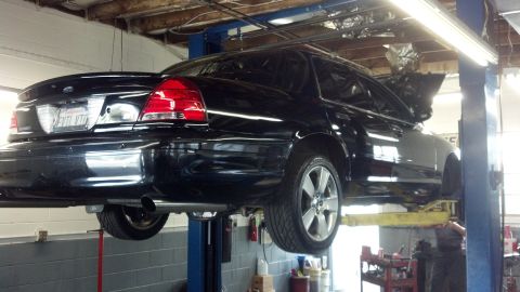 2003 Ford Crown Victoria - Rear view of a black Ford four-door sedan raised on a two-post lift in a repair shop, showing exhaust and silver wheel.
