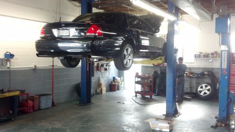 2003 Ford Crown Victoria - black four-door sedan raised on a two-post lift in a shop, with a mechanic at a workbench and tools in the background.