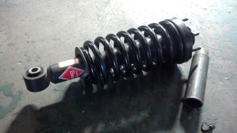 2003 Ford Crown Victoria - strut with black coil spring bearing a red label, lying next to a metal tube on a worn green concrete floor.