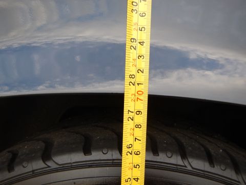 2003 Ford Crown Victoria - yellow tape measure is held against a tire tread and dark wheel arch beneath a cloudy sky.