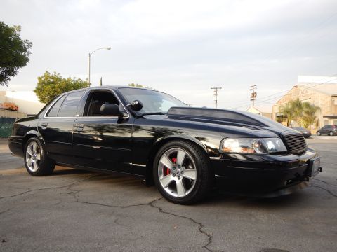 2003 Ford Crown Victoria - lowered black four-door sedan with a mesh grille and silver alloy wheels parked in an industrial lot.
