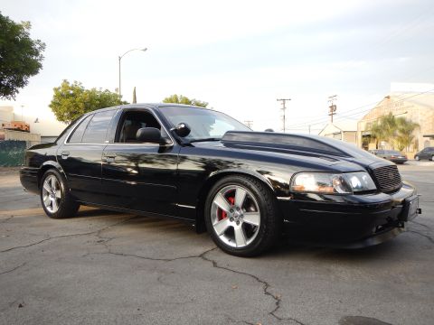 2003 Ford Crown Victoria - glossy black full-size sedan with a mesh grille and silver wheels stands on cracked asphalt in a lot.