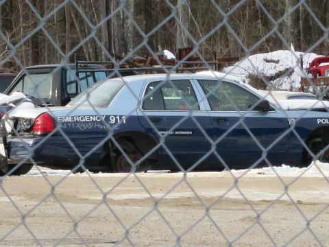 1991 Ford LTD Crown Victoria - A derelict police car sits behind a chain-link fence, showing "EMERGENCY 911" on its side.