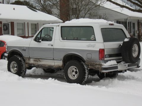 1994 Ford Bronco - A two-tone 1994 Ford Bronco is parked in the snow, showing its rear profile and spare tire.