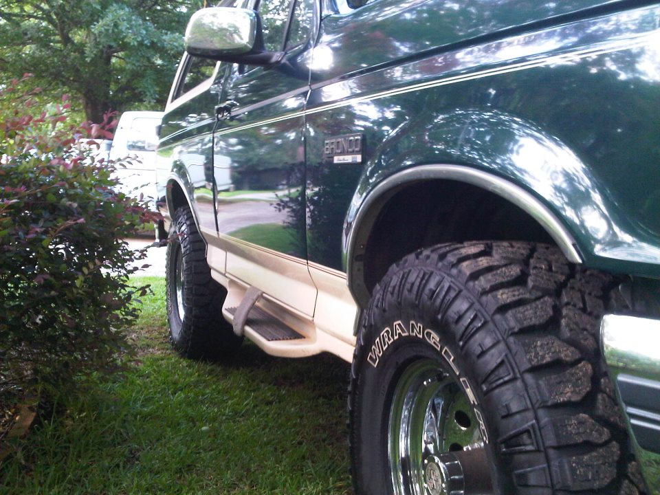 Green 1993 Ford Bronco with tan lower trim and chrome wheels, parked on grass next to bushes