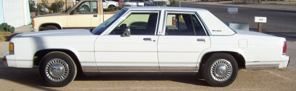 White Ford LTD Crown Victoria sedan, driver-side profile, parked on dirt lot
