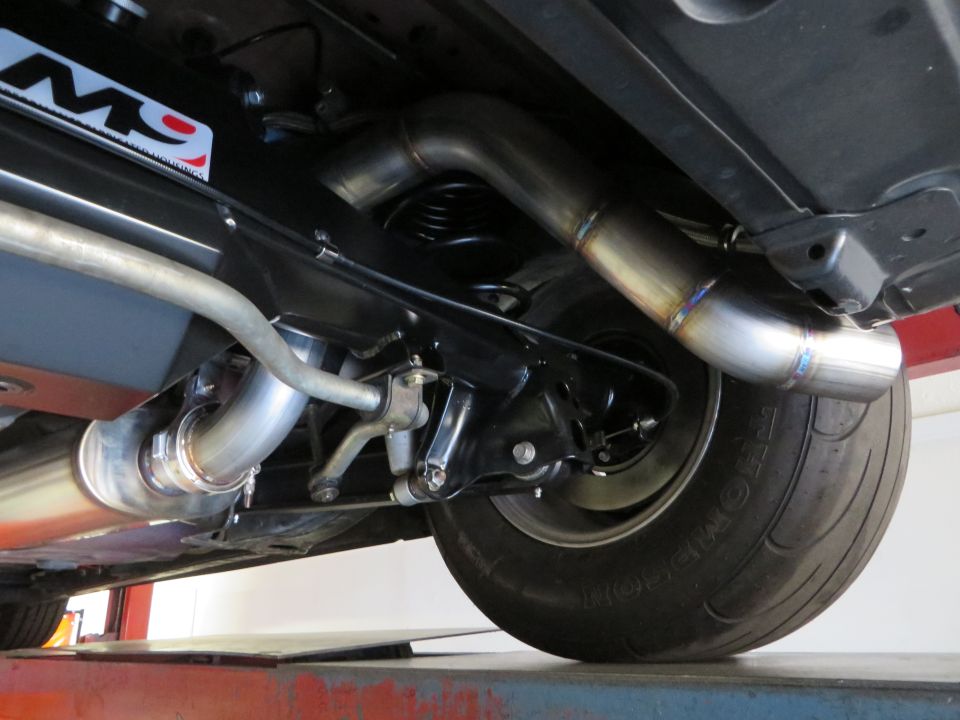 Underside view of a car's rear suspension and custom exhaust system with a "M9" sticker visible