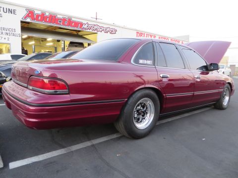 1992 Ford Crown Victoria - A maroon Ford Crown Victoria with drag tires is parked in front of Addiction Motorsports, its hood open.
