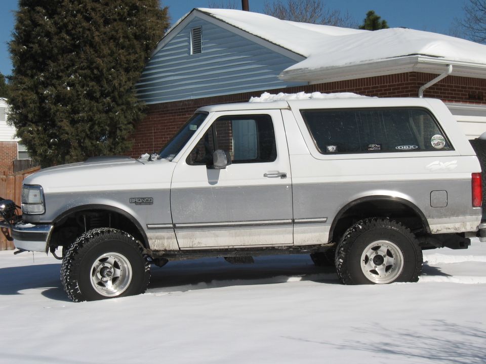 White and silver Ford Bronco parked in snow, side view. Flat front tire