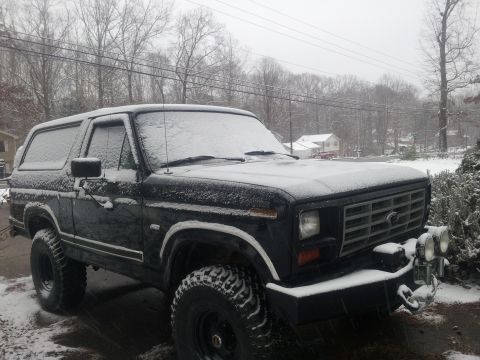 1986 Ford Bronco - A black Ford Bronco sits covered in snow during a winter storm, viewed from the front three-quarter angle.