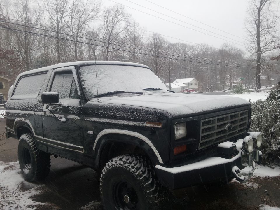 Black Ford Bronco covered in snow, front 3/4 view, with aftermarket lights on the bumper