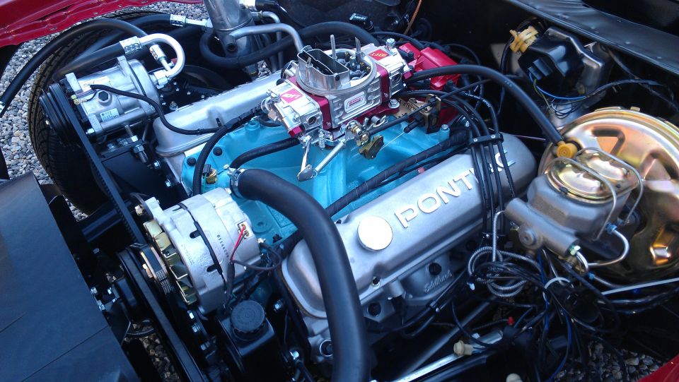 Close-up of a Pontiac V8 engine with blue block and silver valve covers. The 'Pontiac' logo is visible on the valve cover