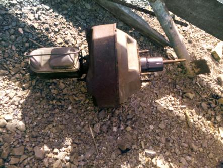 1982 Ford Bronco - This automotive part, likely a brake booster and master cylinder, sits on a bed of gravel with some wooden debris in the background.