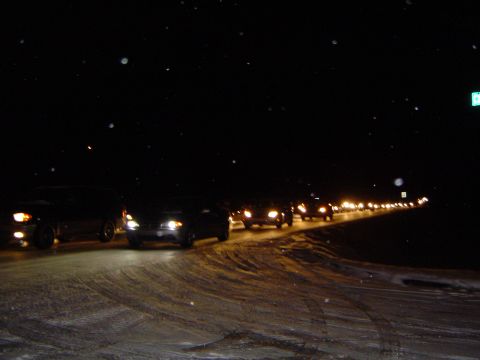 1986 Ford Bronco - Multiple vehicles travel in a convoy down a snow-covered road at night, their headlights cutting through the darkness.