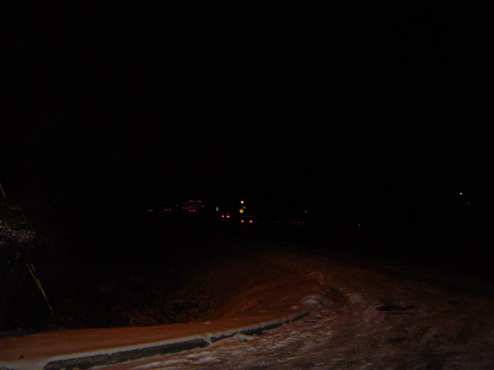 Dark road at night with snow on the side and distant lights visible