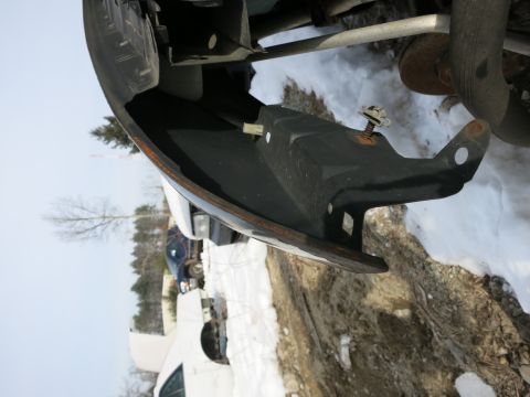 1991 Ford LTD Crown Victoria - Close-up view of a black front bumper component, showing its mounting points and a rusty edge. Other vehicles and snowy ground are visible…