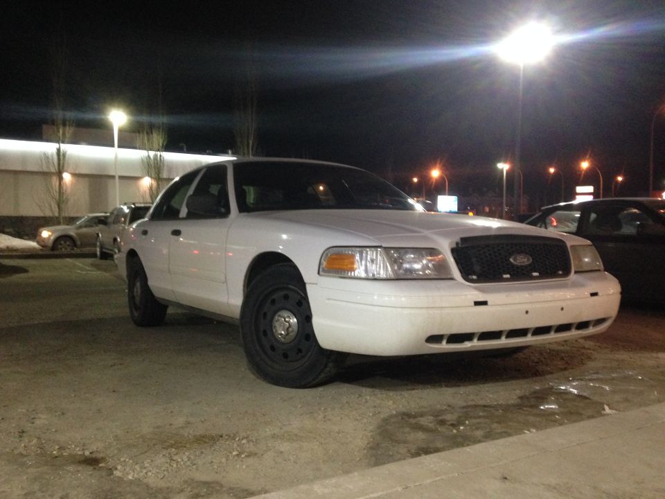 White Ford Crown Victoria with black steel wheels parked on gravel lot at night, front three-quarter view