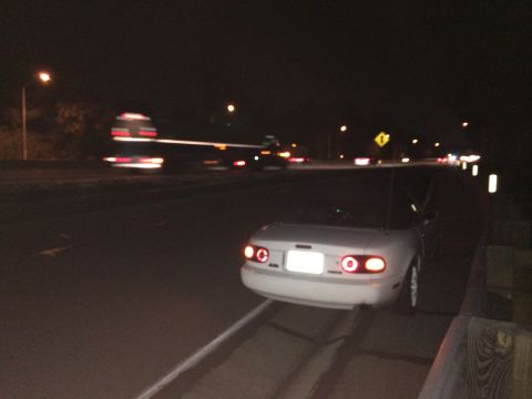 1991 Mazda Miata - A white convertible is parked on the shoulder of a road at night, with traffic passing by.