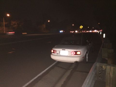 1991 Mazda Miata - A white convertible sports car is parked on the shoulder of a highway at night, with its brake lights illuminated.