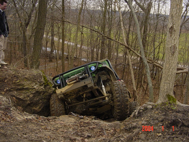 Green off-road vehicle tilted on a muddy incline with trees in the background