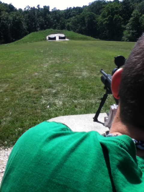 2006 Ford E350 - A person wearing a green shirt and ear protection aims a rifle with a scope at a target on a grassy hill in the distance.