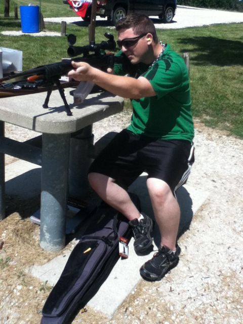 2006 Ford E350 - Man aims rifle over picnic table at outdoor range, gun bag on ground.