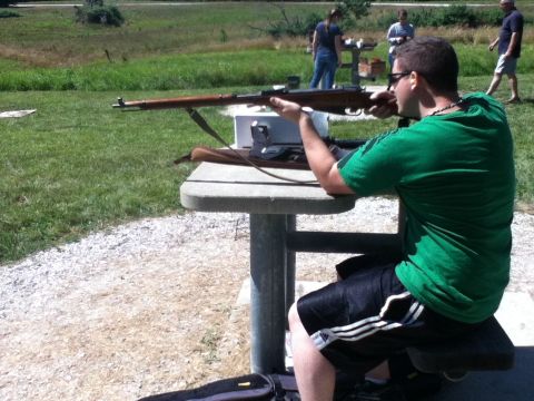 2006 Ford E350 - A man takes aim with a rifle at an outdoor shooting range, with other people present in the background.
