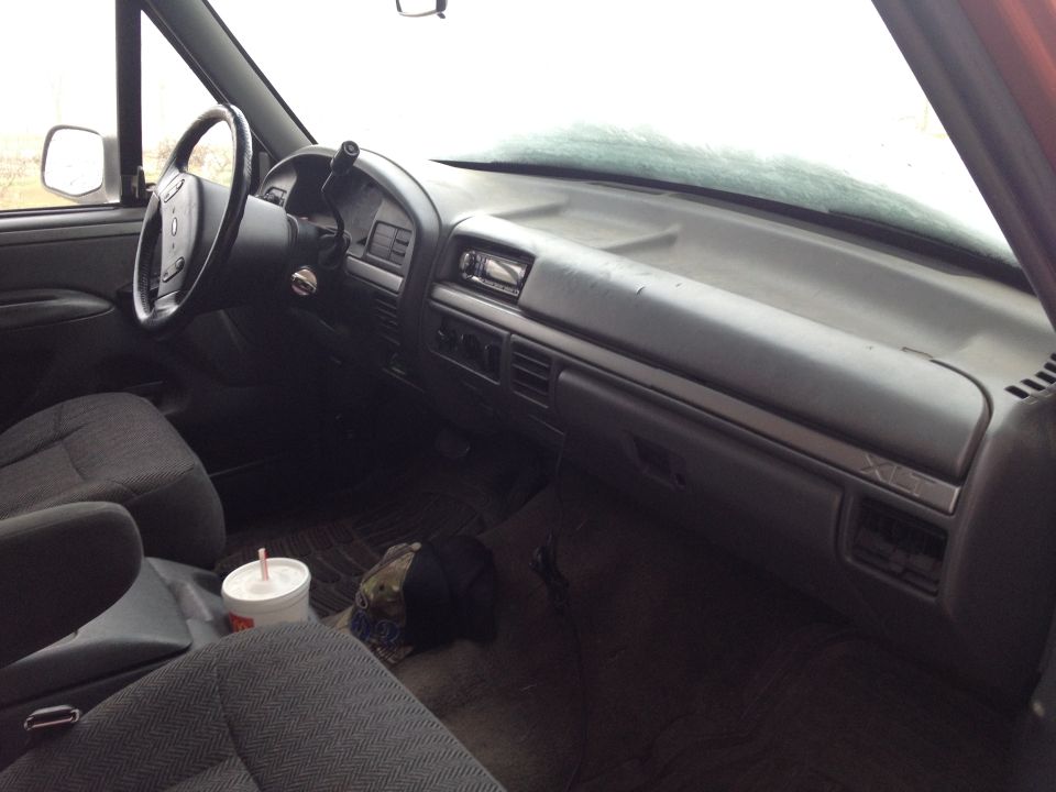 Interior of a truck cab, showing the dashboard, steering wheel, and seats. A cup and a hat are on the floor