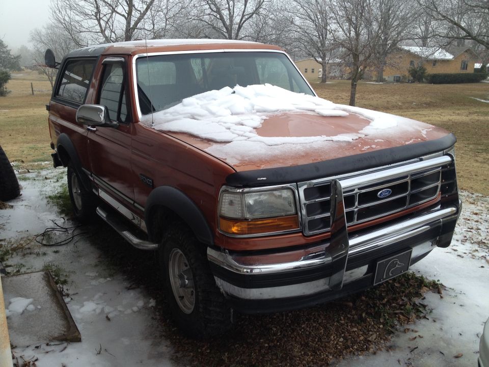 Orange Ford Bronco with snow on the hood and windshield, parked outside in a snowy yard