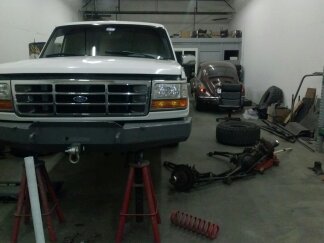 1995 Ford Bronco - A white Ford truck is elevated on jack stands in a garage, with a red Volkswagen Beetle visible in the background.