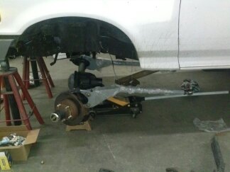 1995 Ford Bronco - The front suspension and frame of a white vehicle are visible while it's elevated for work.