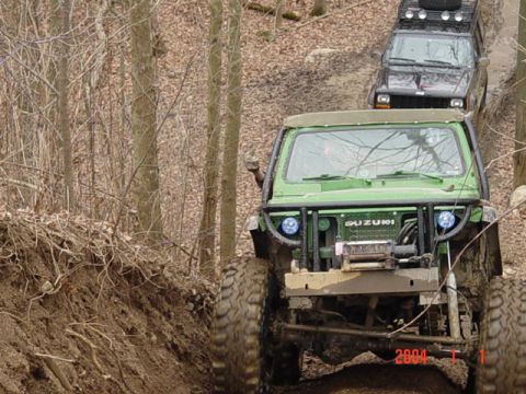 1992 Ford Bronco - Two off-road vehicles navigate a steep, wooded trail covered in mud and fallen leaves.