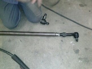 1995 Ford Bronco - Close-up of a vehicle steering linkage rod with ball joints on a concrete floor.