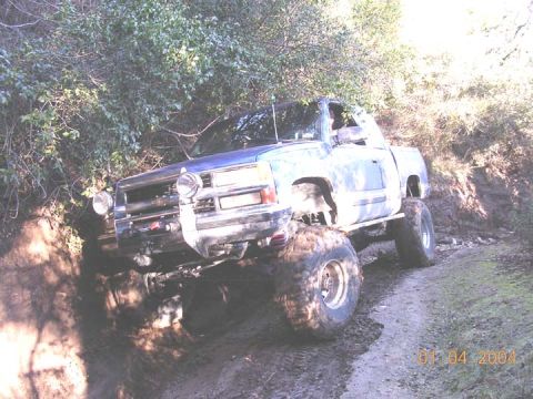 1994 Ford Bronco - A lifted blue pickup truck navigates a challenging, muddy off-road trail.