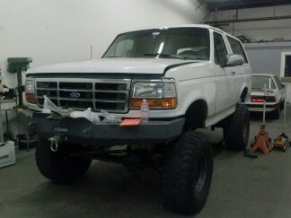 1995 Ford Bronco - Modified white Ford Bronco with large tires and aftermarket bumper, seen in a garage with another vehicle in the background.