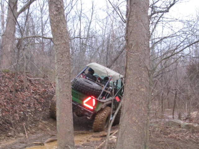 Green off-road vehicle with spare tire on rear, tilted on muddy terrain between trees