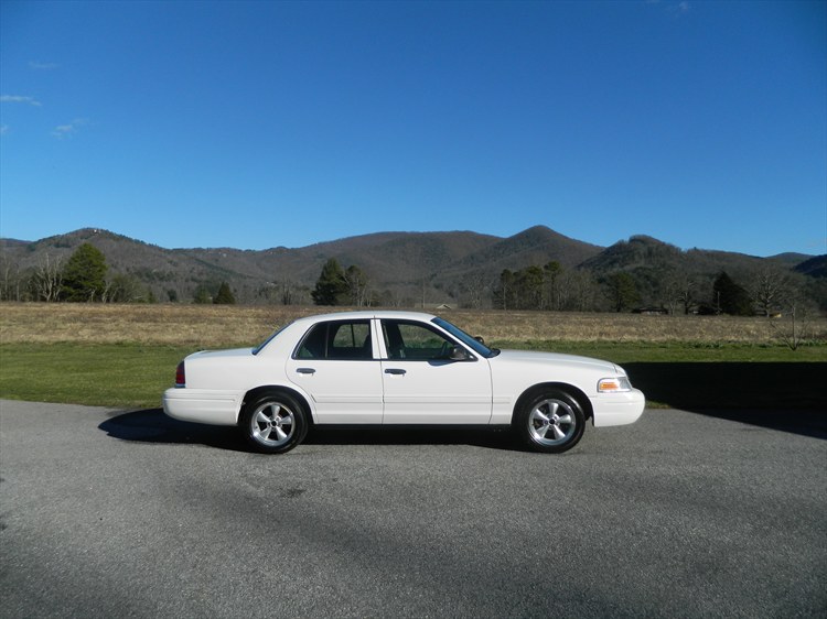White Ford Crown Victoria sedan, side view, parked on road with mountains and blue sky behind