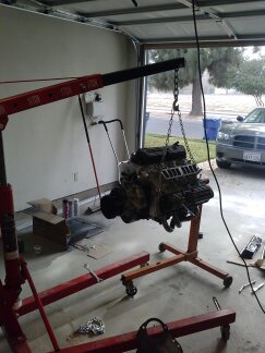 1995 Ford Bronco - An engine is suspended by a red engine hoist in a garage, with a car visible in the background.