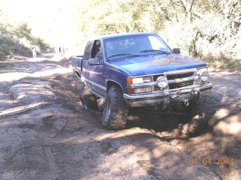 1994 Ford Bronco - A blue pickup truck navigates a challenging muddy terrain with trees in the background.