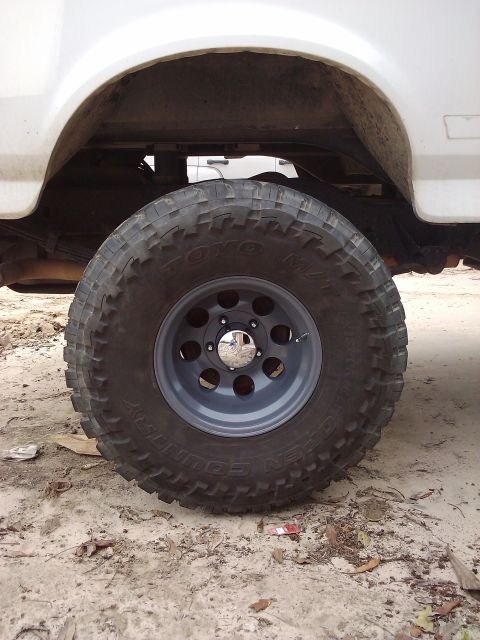 1995 Ford Bronco - A close-up of a large off-road tire and gray rim on a white SUV, with another vehicle visible in the background.