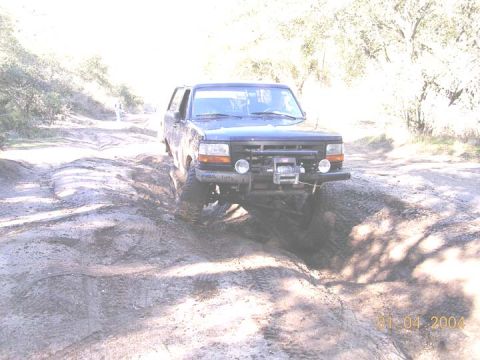 1994 Ford Bronco - Blue 1992-96 Ford Bronco crawling through a dirt rut on a sandy off-road trail, front view in harsh backlit sun. It carries a winch mounted…