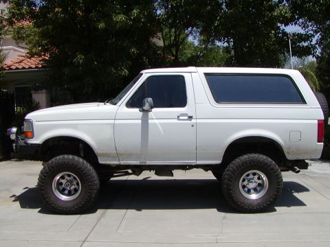 1995 Ford Bronco - A lifted white Ford Bronco is parked on a paved surface with a house and trees in the background.