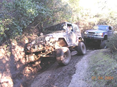 1994 Ford Bronco - A mud-caked Jeep Wrangler with the top off tips hard onto its passenger side climbing out of a rutted trail, front bumper hoop and round dr…